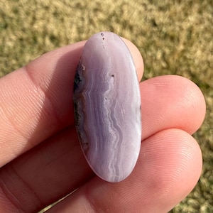 May include: A polished, oval-shaped gemstone with a lavender hue and swirling patterns. The stone has a smooth surface and a mix of light and dark purple tones. Small dark inclusions are visible near the edges. The gemstone is held in a hand.