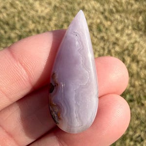 May include: A teardrop-shaped, polished gemstone with a pale lavender hue. The stone features swirling, banded patterns in shades of white and lavender. A small area of brown and orange inclusions is visible near the base. The stone is held in a hand.