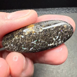 May include: A polished, oval-shaped gemstone with a dark brown base color. The stone features a speckled pattern with iridescent flashes of green, blue, and white. The surface has a slightly textured appearance, with subtle variations in color and tone.