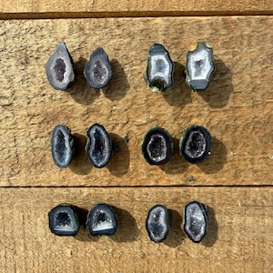 May include: A collection of eleven polished geode slices, each showcasing a unique interior crystal formation. The geodes vary in shape, with a mix of rounded and irregular edges. The outer edges are dark, framing the sparkling crystal centers, set against a wooden background.