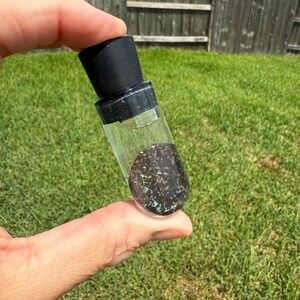 May include: A small, clear glass vial with a black stopper, filled with a clear liquid and a dark, speckled stone. The stone has flecks of blue and green. The vial is held against a background of green grass.