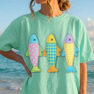 Peut inclure: Un t-shirt à manches courtes vert menthe clair orné de trois motifs de poissons stylisés sur la poitrine. Le poisson de gauche est rose à pois blancs, celui du centre est à carreaux bleus et celui de droite est à vichy jaune, chacun avec un X.