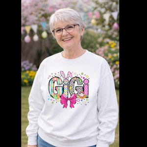 May include: White sweatshirt with the word "Gigi" in colorful, patterned letters. The design includes a pink bow, Easter eggs, and a bunny with a carrot. The sweatshirt is worn by a person with glasses.