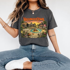 May include: A charcoal gray t-shirt featuring a graphic print with the text HALLOWEENTOWN and SUMMERWEEN EST. 1998. The illustration depicts a tropical pool scene with a jack-o'-lantern in a floatie, palm trees, a skull, and a mansion at sunset.