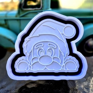 May include: A white plastic cookie cutter shaped like a cartoon Santa Claus wearing a hat, with raised outlines defining the eyes, nose, mustache, and hands resting on the bottom edge, featuring a deep black inner border for contrast.