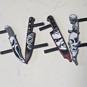 May include: Four decorative drawer pulls shaped like knives, each featuring a unique horror-themed graphic. The designs include a ghost mask, a hockey mask, a doll face, and a skull. Each pull has two black mounting posts on the back.