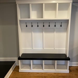 May include: A white wooden entryway organizer with a black bench and hooks. The organizer has open shelves at the top, six black hooks, and cubbies below the bench. The bench is painted black.