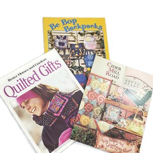 May include: Three quilting pattern books with colourful covers. The first book is titled "Be Bop Backpacks" and features a variety of colourful backpacks. The second book is titled "Better Homes and Gardens Quilted Gifts" and features a woman wearing a pink hat and a colourful scarf. The third book is titled "Cider Mill Road Art-to-Heart" and features a quilt with a red, white, and blue design.