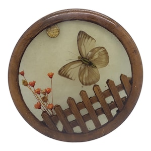 May include: A round wooden frame with a glass insert featuring a brown butterfly with green accents, a small cluster of orange flowers, and a brown wooden fence.