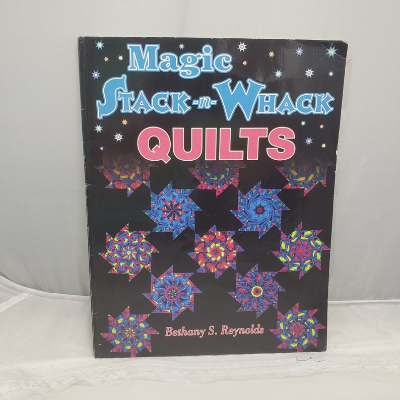 Puede incluir: Portada del libro "Magic Stack-n-Whack Quilts" de Bethany S. Reynolds. La portada presenta un fondo negro con bloques de colcha en forma de estrella de colores.