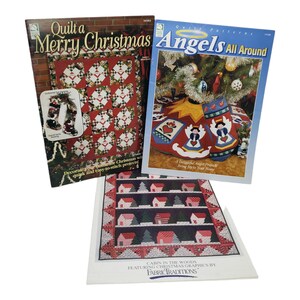 May include: Three quilting pattern books with Christmas themes. The first book is titled "Quilt a Merry Christmas" and features a red and green quilt with a Christmas stocking design. The second book is titled "Angels All Around" and features a quilt with a Christmas tree design. The third book is titled "Cabin in the Woods" and features a quilt with a winter village design.