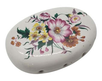 May include: Oval-shaped, white ceramic soap dish with a floral design. The flowers are in shades of pink, yellow, and orange, with green leaves. The dish has several small holes for drainage.
