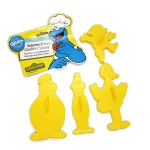 Könnte beinhalten: Satz mit vier gelben, aus Kunststoff gefertigten Ausstechern mit Sesamstraße-Charakteren: Cookie Monster, Big Bird, Elmo und Bert. Die Ausstecher sind auf einer Karte mit dem Sesamstraße-Logo und dem Text "Sesame Street Cookie Cutters" verpackt.