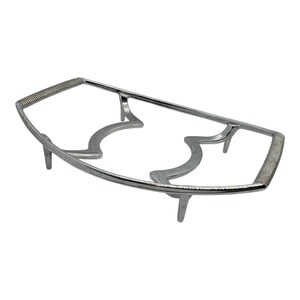 May include: A silver-colored metal trivet with a rectangular frame and decorative curved supports. The handles have a textured, ribbed design. The trivet has four small legs for elevation and is designed to hold hot dishes.