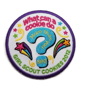 Op de afbeelding: Een witte Girl Scout Cookies-badge met een paarse rand. De badge heeft een blauw vraagteken met de tekst "What can a cookie do? More than you see" en een kleurrijke vallende ster. De tekst "Girl Scout Cookies 2012" staat onderaan de badge.