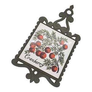 May include: A decorative trivet with a white ceramic tile featuring a cranberry illustration. The tile is framed by a dark green, ornate metal border. The word "Cranberry" is written in cursive on the tile.