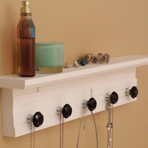 Peut inclure: Étagère en bois blanc avec des boutons en cristal noir et des crochets pour accrocher des colliers. L'étagère a une petite tablette sur le devant et est fixée à un mur. Il y a un bougeoir en verre transparent et une bouteille de liquide sur l'étagère.