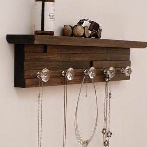 May include: A dark brown wooden shelf with five crystal knobs for hanging necklaces. The shelf has a black painted edge and is mounted on a white wall. A small bottle with a white label is sitting on the shelf.