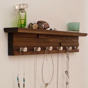 May include: A dark brown wooden wall mounted shelf with a black top and silver hooks. The shelf has a layered design and is decorated with necklaces, bracelets, and a bottle of perfume.