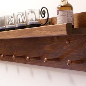 Op de afbeelding: Een donker gebeitst houten plank met zes messing haken. De plank bevat vier glazen kandelaars, een fotolijst en een fles. De plank is aan een witte muur bevestigd.