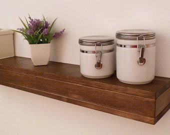 May include: A dark brown wooden shelf with two white ceramic canisters with silver lids and a small potted plant with purple flowers.