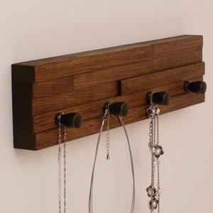 May include: A wooden wall-mounted jewelry organizer with five hooks. The organizer is made of dark brown wood with a black border. There are four necklaces hanging from the hooks.