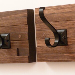 Könnte beinhalten: Zwei Wandhaken aus Holz mit schwarzen Metallhaken. Die Haken sind an einer Wand montiert und haben einen rustikalen, ländlichen Stil.