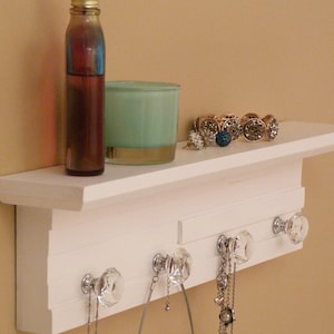 Può includere: Mensola in legno bianco con ganci e pomelli di cristallo. Una bottiglia di liquido, una candela turchese e gioielli sono esposti sullo scaffale.