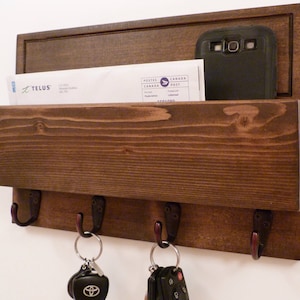 May include: A dark brown wooden wall mounted key holder with a mail slot. The key holder has four hooks and a small shelf for mail. There are two sets of keys and a cell phone on the shelf.