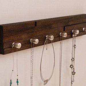 May include: A dark brown wooden wall mounted jewellery organiser with six silver hooks. The organiser has five necklaces hanging from the hooks.