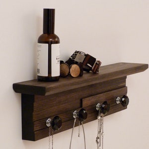 May include: A dark brown wooden shelf with a black trim and five crystal knobs. The shelf has a small ledge on top and is mounted to a white wall. There are necklaces hanging from the knobs and a bottle of liquid and a belt on the ledge.