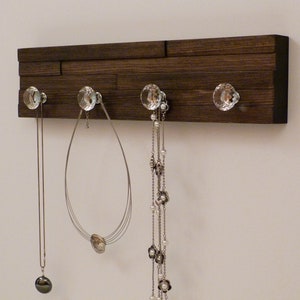 May include: A dark brown wooden wall mounted jewelry organizer with five crystal knobs. Three necklaces hang from the knobs.