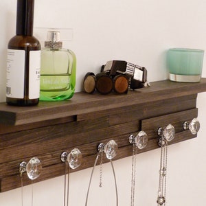 May include: A dark brown wooden shelf with a decorative ledge and five crystal knobs. The shelf has a green glass bottle with the text "Vent de Sud" on it, a clear glass bottle, and a teal glass candle holder. There are necklaces hanging from the knobs.