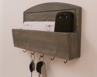 May include: Gray wooden wall mounted key holder with a shelf for mail and a slot for a phone. The key holder has five hooks and is decorated with a distressed gray paint finish.