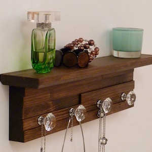 May include: A dark brown wooden shelf with a decorative edge, featuring crystal door knobs as hooks. The shelf is adorned with a green glass bottle of perfume, a brown beaded necklace, and a teal glass candle.