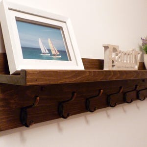 Puede incluir: Un estante de madera teñida oscura con un marco de fotos, un letrero decorativo y una pequeña planta en maceta. Debajo del estante hay seis ganchos de metal para colgar objetos. El marco de fotos contiene una imagen de veleros.