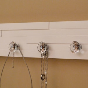 May include: White wooden wall mounted jewellery organiser with four crystal knobs. The organiser has five horizontal shelves and is ideal for hanging necklaces, bracelets, and other jewellery.