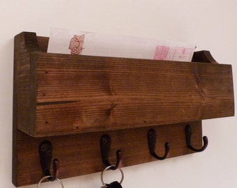 May include: A dark brown wooden wall-mounted organizer featuring a rectangular mail slot compartment at the top and four black metal hooks attached to the bottom panel. A white envelope with a red graphic is tucked inside the wooden mail holder.