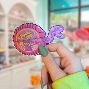 May include: A pink and purple sticker of a Hubba Bubba bubble gum tape dispenser with a pink snake wrapped around it. The sticker says "Hubba Bubba Boa Tape" and "Awesome Original Fun!"