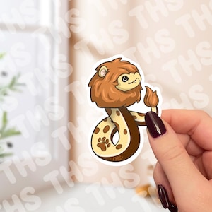 May include: A sticker depicting a cartoon lion-snake hybrid. The lion's mane is brown, and the snake's body is tan with brown spots. The sticker is held by a person with dark nail polish.