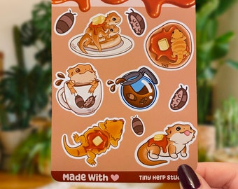 Può includere: Un foglio di adesivi con draghi barbuti dei cartoni animati che si godono la colazione. Il foglio include immagini di un drago che mangia pancake, un drago che beve caffè e un drago che mangia una ciambella. Il foglio è intitolato "Pancake Beardies Sticker Sheet" e include il testo "Made With ♥ Tiny Herp Stuff".
