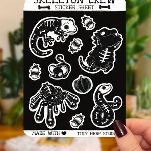May include: A sticker sheet with a black and white design featuring a skeleton crew of various animals, including a gecko, a bearded dragon, a spider, a snake, and a pumpkin. The sheet is titled "Skeleton Crew Sticker Sheet" and includes the text "Made with ♥ Tiny Herp Stud".