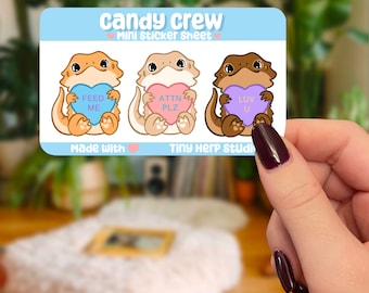 May include: A sticker sheet with three cartoon bearded dragons holding candy hearts. The dragons are brown, orange, and light brown. The hearts say "Feed Me", "Attn Plz", and "Luv U". The sticker sheet is blue and white with the text "Candy Crew Mini Sticker Sheet" and "Made with Tiny Herp Studio".