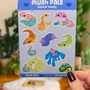 Könnte beinhalten: Eine Sticker-Tafel mit niedlichen Comic-Tieren, darunter eine Schlange, ein Gecko, eine Raupe, eine Schildkröte, eine Spinne und eine Maus. Die Sticker-Tafel trägt den Titel "Plush Pals Sticker Sheet" und hat oben und unten ein Herz-Design. Der Text "Made With" befindet sich unten links auf der Tafel.