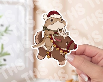 May include: A sticker featuring a cartoon lizard wearing a Santa hat and scarf, riding a reindeer with golden bells. The lizard is light brown, the reindeer is brown, and the hat is red. The sticker is held by a hand.