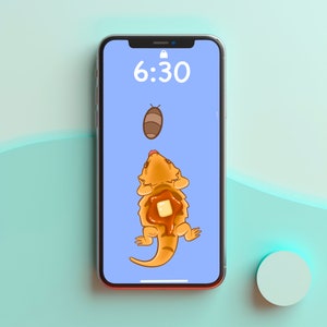 May include: A smartphone screen displaying a digital clock showing 6:30. The background is light blue and features a cartoon image of a bearded dragon with a pancake on its back.