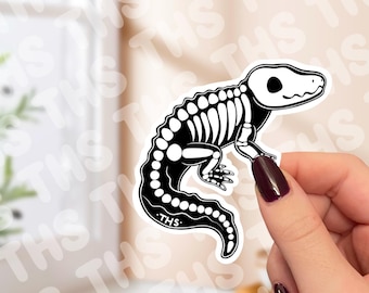 Op de afbeelding: Een zwart-witte sticker met een gestileerde illustratie van een hagedisskelet. De sticker heeft een afgeronde vorm en de tekst "THS" is zichtbaar. De hagedis wordt in een zijaanzicht weergegeven.