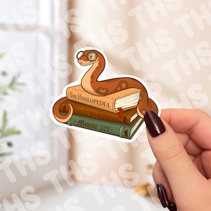 May include: A sticker featuring a cartoon snake wearing glasses, perched atop a stack of books. The books are titled "The Hisslopedia", "How To Speak Human", and "Mouse 101". The snake is brown with a light brown belly.