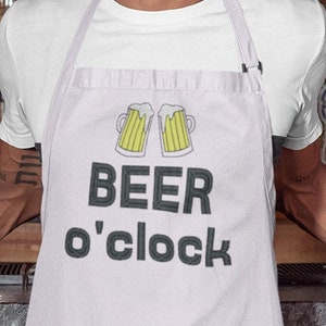 Może przedstawiać: Biały fartuch z jasnofioletowym wykończeniem i wzorem dwóch żółtych kufli piwa z napisem "BEER o'clock".