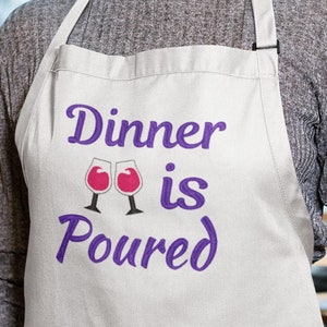 May include: White apron with purple embroidery that reads "Dinner is Poured" with two wine glasses. The embroidery is in a cursive font. A woman is wearing the apron.